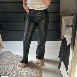 Svarta skinnbyxor - Snygga svarta skinnbyxor med hög midja och straight passform. Byxorna har en stilren design med dragkedja och knapp framtill. Perfekta för en trendig look. Defekt på höften, syns inte super mycker och går att laga själv eller hos sömmerska, därav billigt pris