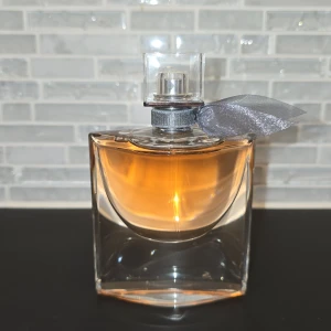 La Vie Est Belle Eau de Parfum - Säljer en elegant flaska av La Vie Est Belle Eau de Parfum från Lancôme. Flaskan har en vacker design med en grå rosett runt halsen och en varm bärnstensfärgad vätska. Perfekt för den som älskar en sofistikerad och tidlös doft.