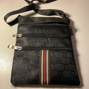 Svart axelväska med mönster - Snygg svart axelväska med diskret mönster och tre dragkedjeförsedda fack. Väskan har en justerbar axelrem och en stilren randdetalj i rött, grönt och beige. Perfekt för att hålla ordning på dina saker med stil.