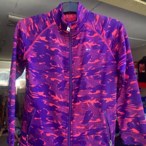 Lila och rosa träningsjacka från Mywear Active - Säljer en cool träningsjacka från Mywear Active i lila och rosa med ett unikt mönster. Jackan har dragkedja och är perfekt för träning eller en aktiv vardag. Den är långärmad och har en sportig look. Perfekt för dig som vill sticka ut!