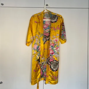 Gul kimono med blommigt mönster - Säljer en superfin gul kimono med ett färgglatt blommigt mönster. Den är i ett glansigt material som känns lyxigt och har trekvartsärmar. Perfekt för sommarens alla fester eller bara för att känna sig extra glam hemma! 💛