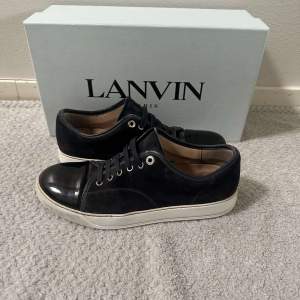 Säljer ett par snygga mörkblå sneakers från Lanvin. De har en stilren design med en blank tå och snörning framtill. Skorna har en vit sula som ger en cool kontrast till det svarta. Perfekta för både vardag och fest!