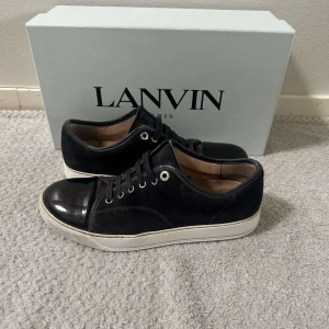 Mörkblå  Lanvin - Säljer ett par snygga mörkblå sneakers från Lanvin. De har en stilren design med en blank tå och snörning framtill. Skorna har en vit sula som ger en cool kontrast till det svarta. Perfekta för både vardag och fest!