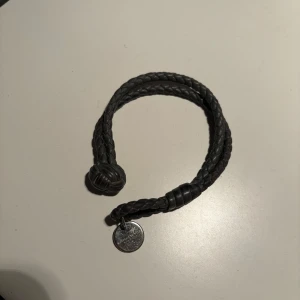 Svart flätat armband med berlock - Säljer ett snyggt svart flätat armband från Bottega. Armbandet har en cool knutdetalj som lås. Perfekt accessoar för både vardag och fest!