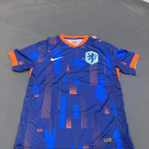 Säljer en snygg blå fotbollströja från Nike med orange detaljer. Tröjan har korta ärmar och ett coolt mönster i blått och orange. Den har KNVB-loggan på bröstet och är i nyskick. Perfekt för fotbollsfans!