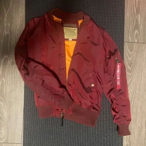 Vinröd bomberjacka från Alpha Industries - Säljer en snygg vinröd bomberjacka från Alpha Industries. Den har en cool orange insida och en klassisk dragkedja framtill. Jackan är långärmad och har en ficka med dragkedja på ärmen. Perfekt för höst och vår!
