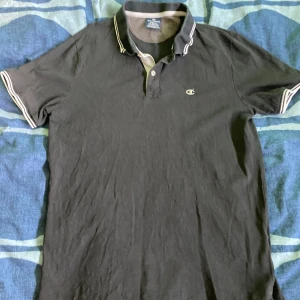 Svart pikétröja från Champion - Säljer en klassisk svart pikétröja från Champion. Den har en snygg krage med vita detaljer och korta ärmar med matchande vita ränder. Perfekt för en avslappnad men stilren look!