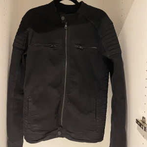 Svart jacka med dragkedja - Säljer en snygg svart jacka med coola detaljer. Den har en dragkedja framtill och två bröstfickor med dragkedjor. Ärmarna är långärmade och har en quiltad design på axlarna och sidorna. Perfekt för höst och vår!