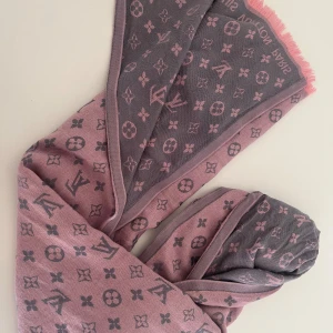 Grå och rosa halsduk från Louis Vuitton - Säljer en superfin grå och rosa halsduk från Louis Vuitton. Den har ett klassiskt monogrammönster och är perfekt för att ge lite lyx till din outfit. Halsduken är mjuk och skön, perfekt för kyliga dagar. Den har fransar i kanterna som ger en extra touch av stil.