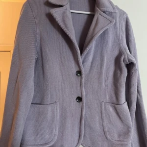 Ljuslila kavaj från Anna Holtblad - Säljer en superfin ljuslila kavaj från Anna Holtblad. Den är stickad och har en snygg krage med två knappar framtill. Perfekt för både vår och höst! Kavajen har långa ärmar och två praktiska fickor. Den är i storlek XL och passar perfekt för en chic och bekväm look. Den har lite noppor som lätt kan tas bort med en borste 