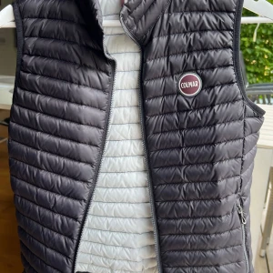 Svart dunväst från Colmar - Säljer en snygg och praktisk svart dunväst från Colmar. Västen har en dragkedja framtill och är perfekt för kyligare dagar. Den är lätt och bekväm att bära, vilket gör den idealisk för både vardag och utomhusaktiviteter. Passar till en person somr 150-160cm