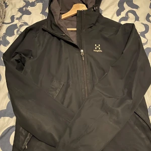Svart regnjacka från Haglöfs - Säljer en svart vindjacka från Haglöfs. Den har en praktisk dragkedja framtill och justerbar huva. Jackan är perfekt för regniga dagar och har en stilren design. Insidan är fodrad med mesh för extra komfort. Den är ganska använd men har inte några skador eller så