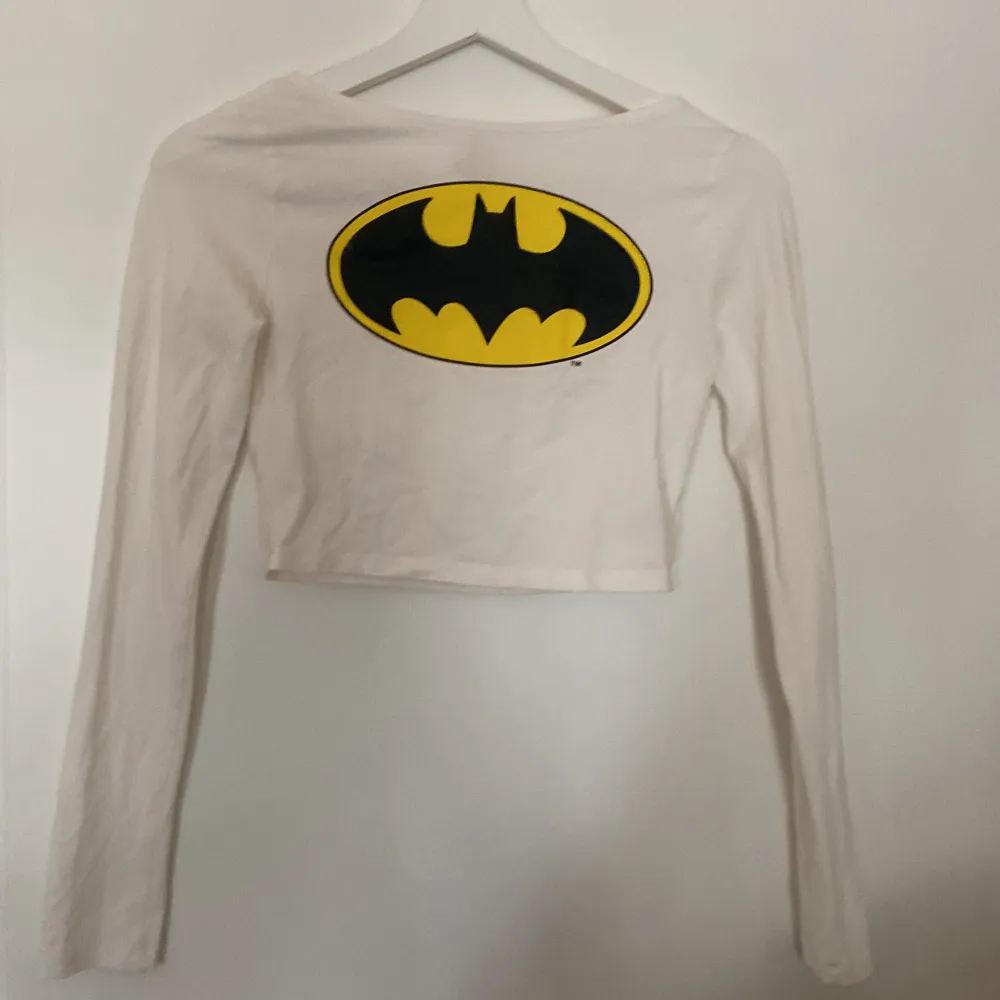 Säljer en supercool vit croppad topp från H&M med den ikoniska Batman-loggan i svart och gult på framsidan. Toppen har långa ärmar och är perfekt för en avslappnad och stilren look. Perfekt för alla Batman-fans!. Toppar.