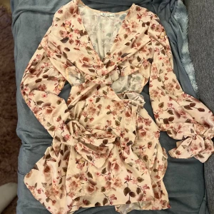 Blommig klänning från NAKD - Säljer en superfin blommig klänning. Den har en v-ringad design med långa ärmar och ett knytband i midjan. Perfekt för vår och sommar! 🌸