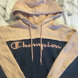 Beige och blå hoodie från Champion - Säljer en snygg och bekväm hoodie från Champion i rosa och blått. Den har en stor logga på bröstet och en klassisk känguruficka. Perfekt för en avslappnad stil eller för att hålla sig varm under kyliga dagar. 🧢