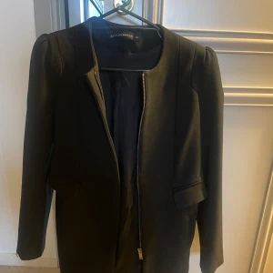 Svart kappa från Zara Woman - Snygg svart kappa från Zara Woman i storlek M. Den har en stilren design med dragkedja framtill och långa ärmar. Perfekt för höst och vår, och funkar både till vardags och fest. Kappan är i bra skick och ger en elegant look.