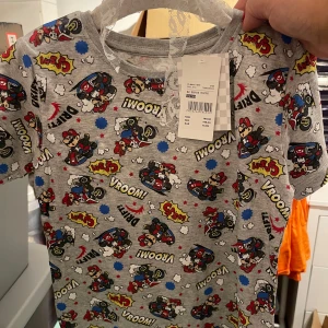 Grå t-shirt med  super mario tryck - Supercool grå t-shirt med färgglatt tryck av bilar och text som 'Vroom!' och 'Wow!'. Perfekt för små bilälskare! T-shirten är kortärmad och gjord i mjuk bomull, vilket gör den bekväm för lek och äventyr. Passar perfekt till vardags eller lekstunder.