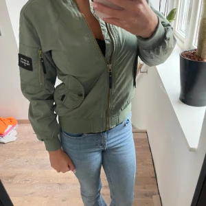 Grön bomberjacka - Säljer en snygg grön bomberjacka jag skulle säga i storlek XXS, Jackan har dragkedja och ribbade muddar vid ärmslut och nederkant. Perfekt för höst och vår! Den har en cool patch på ärmen och fickor med knappar framtill. Passar både till vardags och fest. Skriv för fler bilder eller frågor ❤️