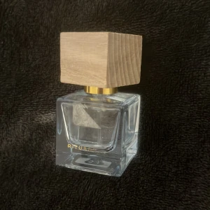 Parfymflaska med trälock - Säljer en stilren parfymflaska med ett fyrkantigt trälock och en elegant glasflaska. Flaskan har en minimalistisk design med guldiga detaljer och märket 'Rituals' på framsidan. Perfekt för den som gillar en naturlig och sofistikerad look.