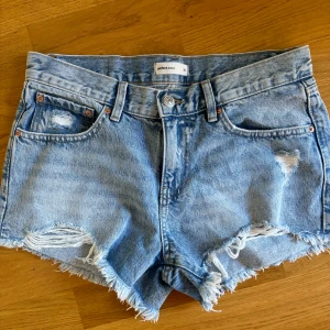 Blå jeansshorts från Gina  - Säljer ett par snygga blå jeansshorts från Gina. De har en cool distressed look med slitningar och fransar vid benen. Perfekta för sommaren och festivaler! De är lågmidjade och har en klassisk femficksdesign. Passar perfekt till en avslappnad stil.