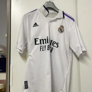 Säljer en vit Real Madrid fotbollströja. storlek L men passar M.Tröjan har korta ärmar och är i Climacool-material för bra ventilation. Den har Real Madrids klubbmärke på bröstet och Benzema 9 tryckt på ryggen. Perfekt för alla fans av laget eller Be