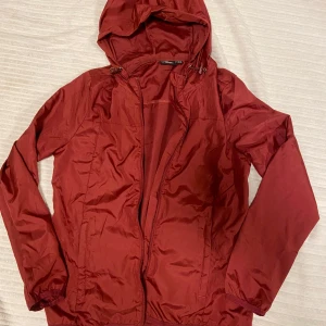 Vinröd löparjacka från Crane - Säljer en röd vindjacka/löparjacka från Crane med huva och dragkedja. Lätt att bära med sig, dragkedja i fickorna ✨