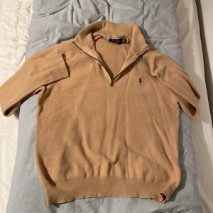 Beige tröja med dragkedja från Ralph Lauren - Säljer en stilren beige tröja från Ralph Lauren med halv dragkedja och broderad logga på bröstet. Tröjan har långa ärmar och ribbade muddar vid ärmslut och nederkant. Perfekt för en avslappnad men snygg look.