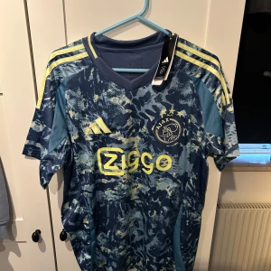 Blå Ajax fotbollströja från Adidas - Snygg blå Ajax fotbollströja från Adidas med ett unikt mönster i olika blå nyanser och gula detaljer. Tröjan har korta ärmar och klubbens logga samt sponsortryck på framsidan. Perfekt för alla Ajax-fans!