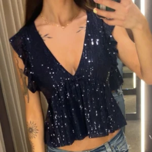 Marinblå glittrig topp - Snygg blå glittrig topp med v-ringning och volangdetaljer. Perfekt för en kväll ute eller fest. Toppen har en lös passform. 
