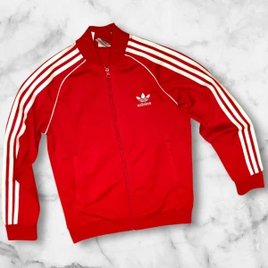 Adidas Originals SST Track Top - Säljer en klassisk röd träningsjacka från Adidas med vita ränder längs ärmarna och dragkedja framtill. Jackan har en broderad Adidas-logga på bröstet och långa ärmar. Perfekt för sportiga tillfällen eller en avslappnad stil.