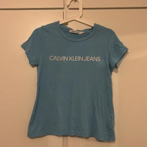 Blå t-shirt från Calvin Klein Jeans - Snygg blå t-shirt från Calvin Klein Jeans med vit logga på bröstet. Den är kortärmad och gjord i mjuk bomull, perfekt för en avslappnad stil. Passar bra till jeans eller shorts för en cool look.          Den några små fläckar vid varandra men som jag tycker inte syns så mycket. Om man tittar på sista och första bilden så syns det inte mycket alls❣️