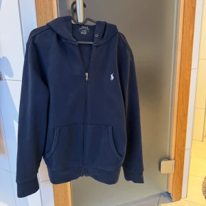 Mörkblå hoodie från Polo Ralph Lauren - Säljer en stilren mörkblå hoodie från Polo Ralph Lauren med dragkedja och klassisk logga på bröstet. Tröjan har långa ärmar och en praktisk huva. Perfekt för en avslappnad look. Obs Ända defekten är att snörerna ej med följer!