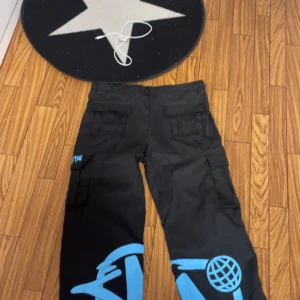 Svarta cargopants minus two  - Coola svarta cargopants med blå graffiti-detaljer. Byxorna har flera fickor och en avslappnad passform. Perfekta för en streetwear-look.