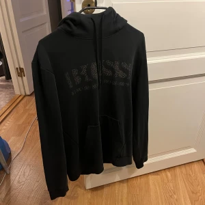 Svart hoodie från Boss - Snygg svart hoodie från Boss med logotyp i guld på framsidan. Tröjan har en klassisk design med huva och samt en stor ficka framtill. Perfekt för en stilren och bekväm look. Men ett litet håll i fickan