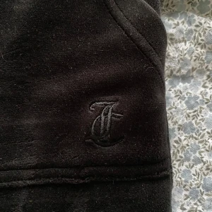 Svarta mjukisbyxor från Juicy Couture - Snygga svarta mjukisbyxor från Juicy Couture med broderad logga. Byxorna har elastisk midja med snörning och praktiska bakfickor med knappar. Perfekta för en avslappnad stil.