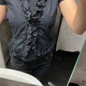 Svart blus med volangdetaljer - Snygg svart blus med volangdetaljer längs framsidan och nederkanten. Perfekt för en stilren look. Kortärmad och i ett mjukt material som ger en bekväm passform.