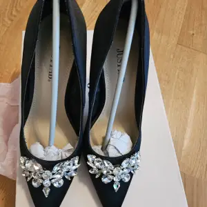 Eleganta svarta pumps från JustFab med spetsig tå och glittrande strassdetaljer framtill. Skorna har en hög klack och är perfekta för att ge en lyxig touch till din outfit.