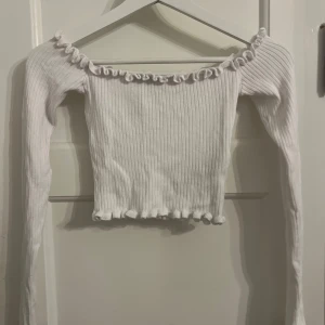 Vit ribbad topp - Säljer en söt vit ribbad topp med långa ärmar och volangdetaljer vid halsringningen och nederkanten. Perfekt för en stilren och trendig look.