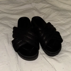 Svarta sandaler med korsade remmar - Snygga svarta sandaler med korsade remmar i ett glittrigt material. Perfekta för en avslappnad stil med en touch av glamour. Sandalerna har en platt sula och öppen tå. Priset kan diskuteras!!