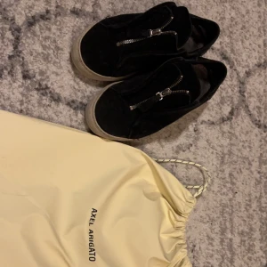 Svarta sneakers från Axel Arigato - Snygga svarta sneakers från Axel Arigato med dragkedja framtill. Skorna har en robust sula och kommer med en praktisk förvaringspåse. Perfekta för en stilren look.