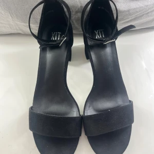 Svarta sandaler med klack från XIT - Snygga svarta sandaler från XIT med en stilren design. De har en bred rem över tårna och en justerbar rem runt ankeln. Klacken är blockformad och ger ett stabilt stöd. Perfekta för en elegant look. Endast använda vid 1 tillfälle.