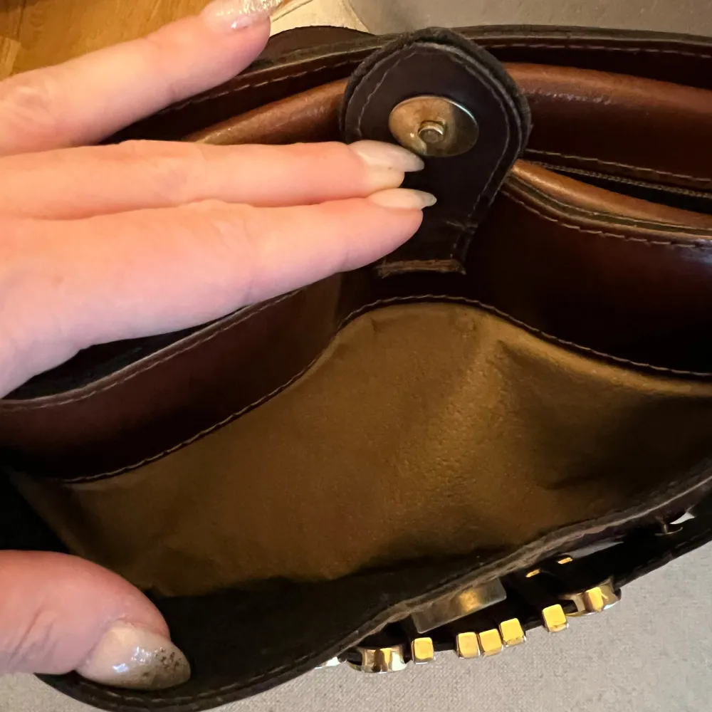 100% äkta Moschino vintageväska - vintage bag. Köptes för ca 15 år sedan som vintage så kvitto finns ej men 100% äkta. Moschino i gulddetaljer. Rödbrunt läder. Lång axelrem. Ytterficka som stängs med knapp och liten innerficka som stängs med dragkedja. Mått ca: 25cm x 17cm. Hämtas eller postas med spårbarhet.. Laukut & Käsilaukut.