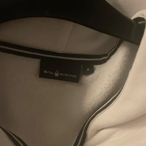 Vit hoodie med dragkedja från Sail Racing - Säljer en stilren vit jacka från Sail Racing med dragkedja och huva. Jackan har en broderad logga på bröstet och praktiska fickor framtill. 
