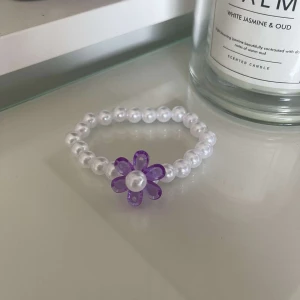 Pärlarmband med lila blomma - Säljer ett supergulligt armband med vita pärlor och en lila blomma i mitten. Perfekt för att piffa upp vilken outfit som helst! Armbandet är elastiskt och passar de flesta storlekar. 🌸
