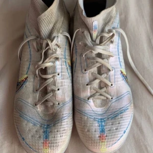 Nike fotbollsskor med färgglada detaljer - Säljer ett par Nike fotbollsskor i vitt med coola färgglada detaljer. Skorna har en tight passform och är perfekta för fotbollsträning. De har snörning och en stickad del vid ankeln för extra stöd. Sulan har en grym design med blå och gula inslag.