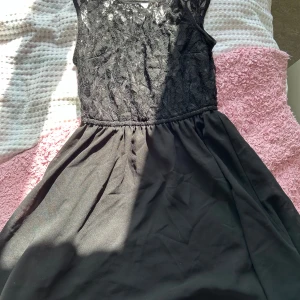 Svart klänning med spetsdetaljer - Säljer en elegant svart klänning med vacker spets upptill. Klänningen har en tight passform upptill och en utsvängd kjol. Perfekt för festliga tillfällen!