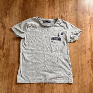 Grå t-shirt från Lager 157 - Säljer en grå t-shirt från Lager 157 i storlek S. Den är gjord av 95% bomull och 5% viskos, vilket gör den supermjuk och skön att ha på sig. T-shirten har ett tryck på framsidan med en byggnad och texten '157'. Perfekt för en avslappnad stil!
