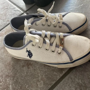 Vita sneakers från U.S. Polo Assn. - Säljer ett par vita sneakers från U.S. Polo Assn. med blå detaljer och snörning. Skorna har en klassisk design med logga på sidan och metallspänne på snörena. Perfekta för en avslappnad stil!