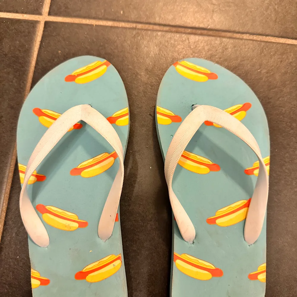 Säljer ett par roliga flip-flops i blått med ett färgglatt korvmönster. Perfekta för sommaren och strandhäng! De har vita remmar och är i bra skick. Storlek 36/37.. Skor.