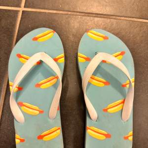 Säljer ett par roliga flip-flops i blått med ett färgglatt korvmönster. Perfekta för sommaren och strandhäng! De har vita remmar och är i bra skick. Storlek 36/37.